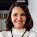 Smiling woman with shoulder length dark hair wearing a white turtleneck sweater indoors in front of a bookshelf