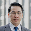 Portrait of a man with short dark hair and glasses wearing a gray suit and blue tie outdoors near a blurred building background